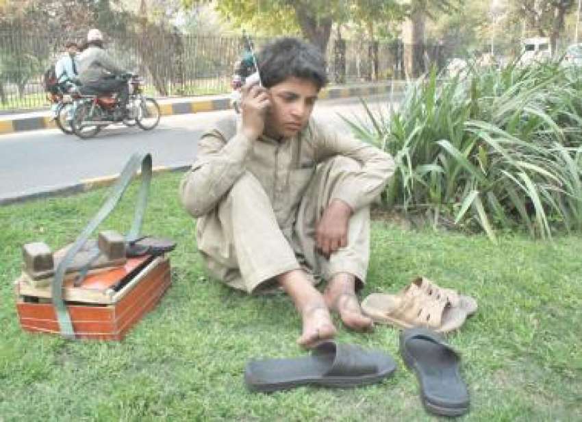 لاہور، ایک محنت کش بچہ گرین بیلٹ پر بیٹھا اپنے عزیز سے موبائل ..